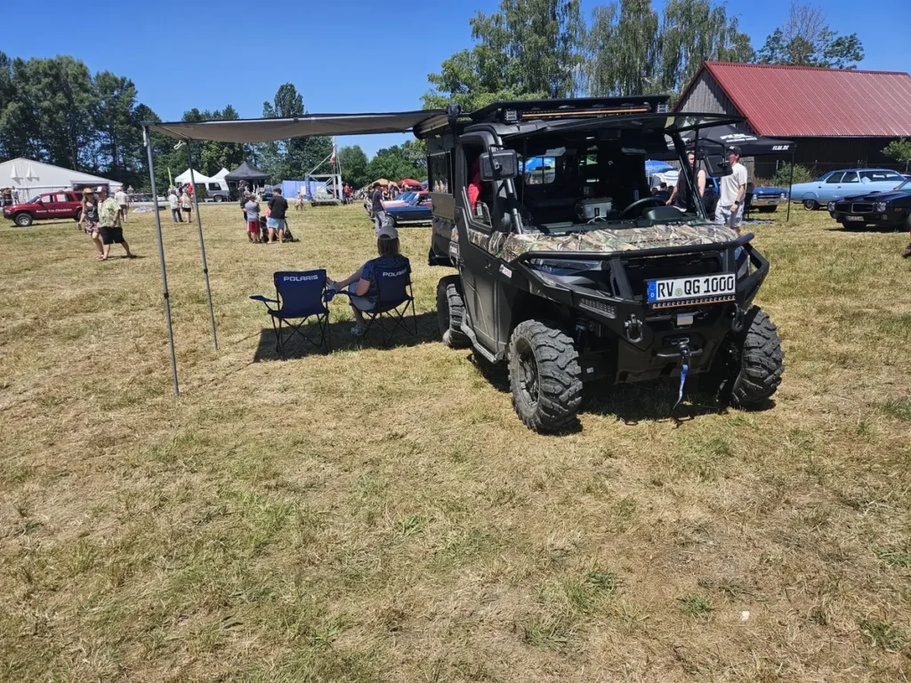 Ein schwarzes Polaris UTV mit Tarnmuster, Expeditionsausrüstung und ausgefahrener Markise auf einer Wiese.