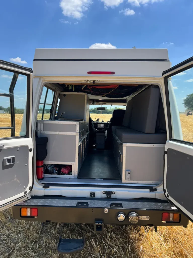Heckansicht eines weißen Overlanding-Fahrzeugs mit Selbstausbau, geöffneten Hecktüren und Aufstelldach in einem Feld.