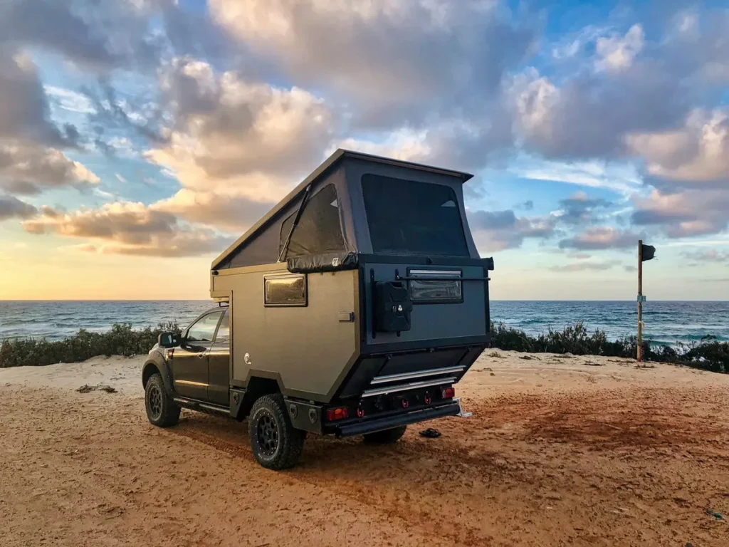 Dunkler 4x4 Pick-up mit Pop-top Wohnkabine am sandigen Strand bei Sonnenuntergang.