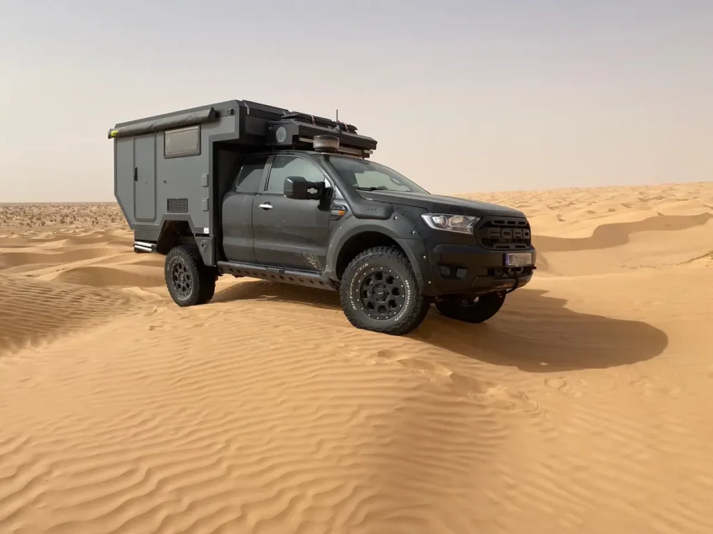 Dunkelgraues Expeditionsmobil mit Pick-up Wohnkabine und Markise in einer Sanddünenlandschaft.