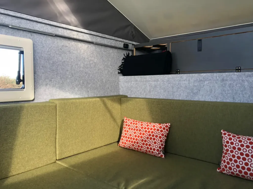 Campervan-Interieur mit olivgrüner Sitzgruppe, rotem Kissen und grauer Filzverkleidung.