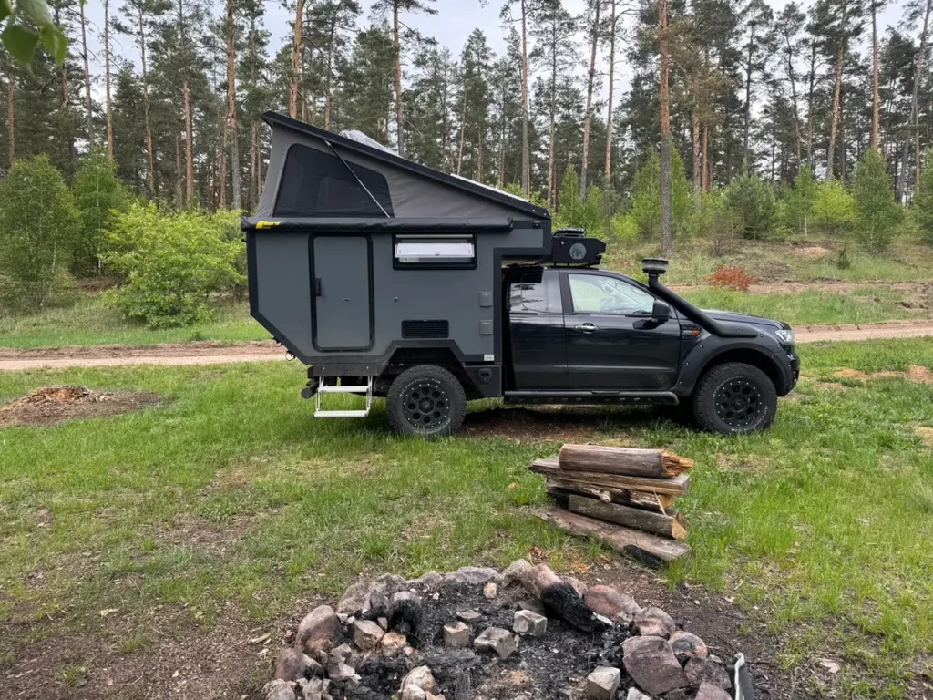 Ein Expeditionsmobil Pick-up mit Pop-top Wohnkabine parkt im Wald neben einer Feuerstelle.